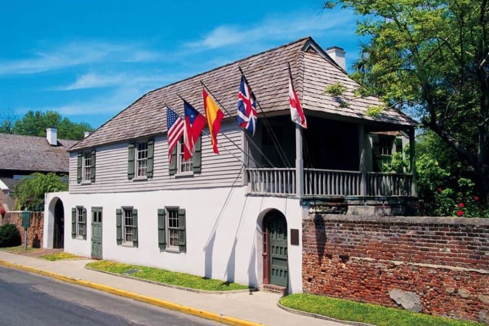 A historic building with multiple flags displayed outside, featuring green shutters and a stone wall.
