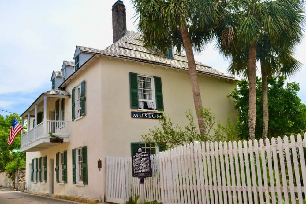A historic museum building with green shutters, surrounded by palm trees and a picket fence.