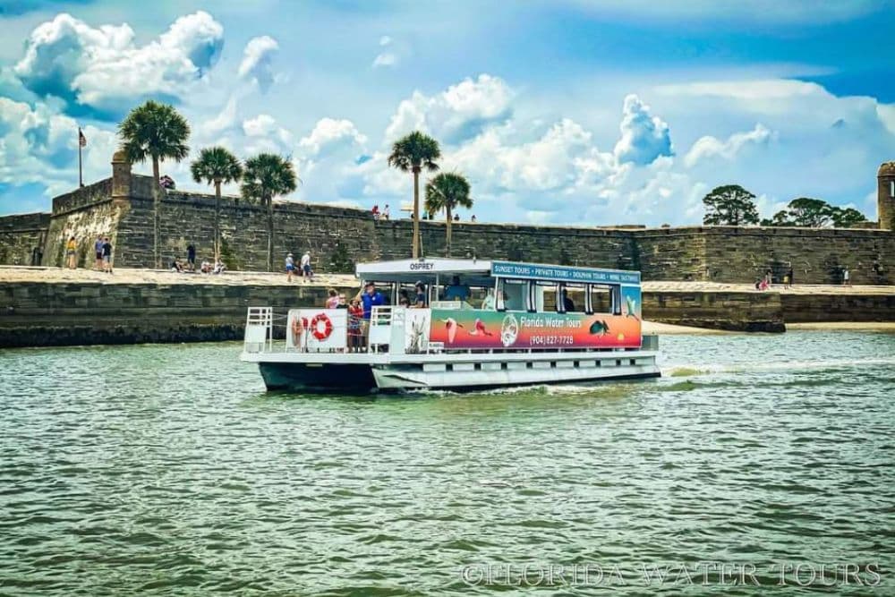 A tour boat named "Osprey" glides through the water near a historic stone fort under a cloudy sky.