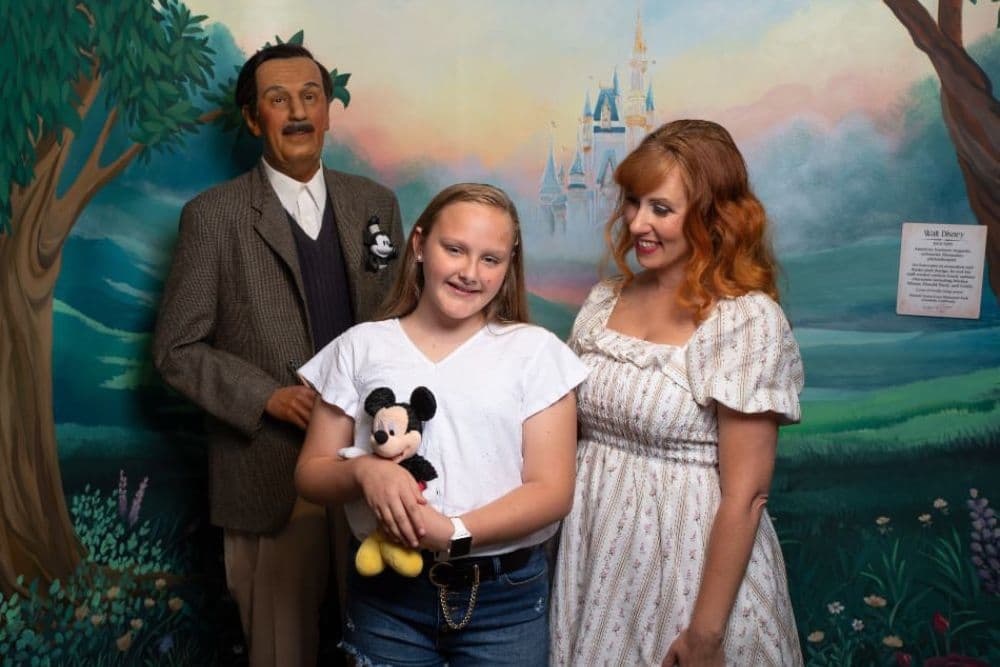 A girl holds a stuffed Mickey Mouse while posing with a woman and a wax figure of Walt Disney in front of a painted backdrop.