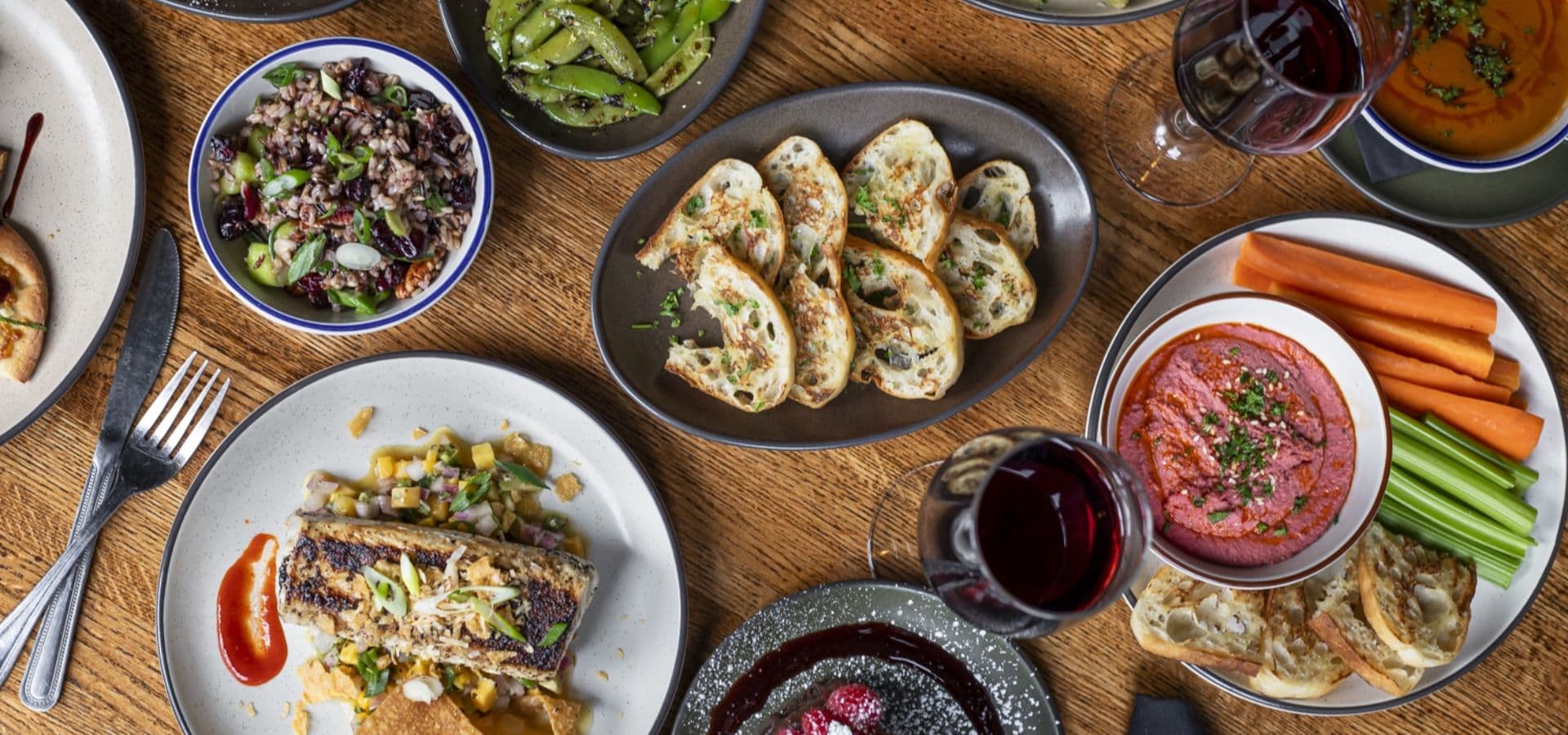 A variety of colorful dishes, including salad, roasted vegetables, and dip, are arranged on a wooden table with glasses of red wine.