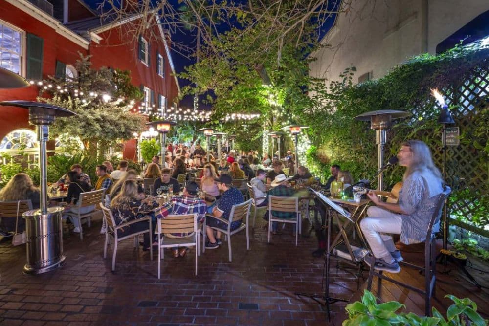 A lively outdoor dining scene at night with musicians and diners surrounded by greenery and string lights.