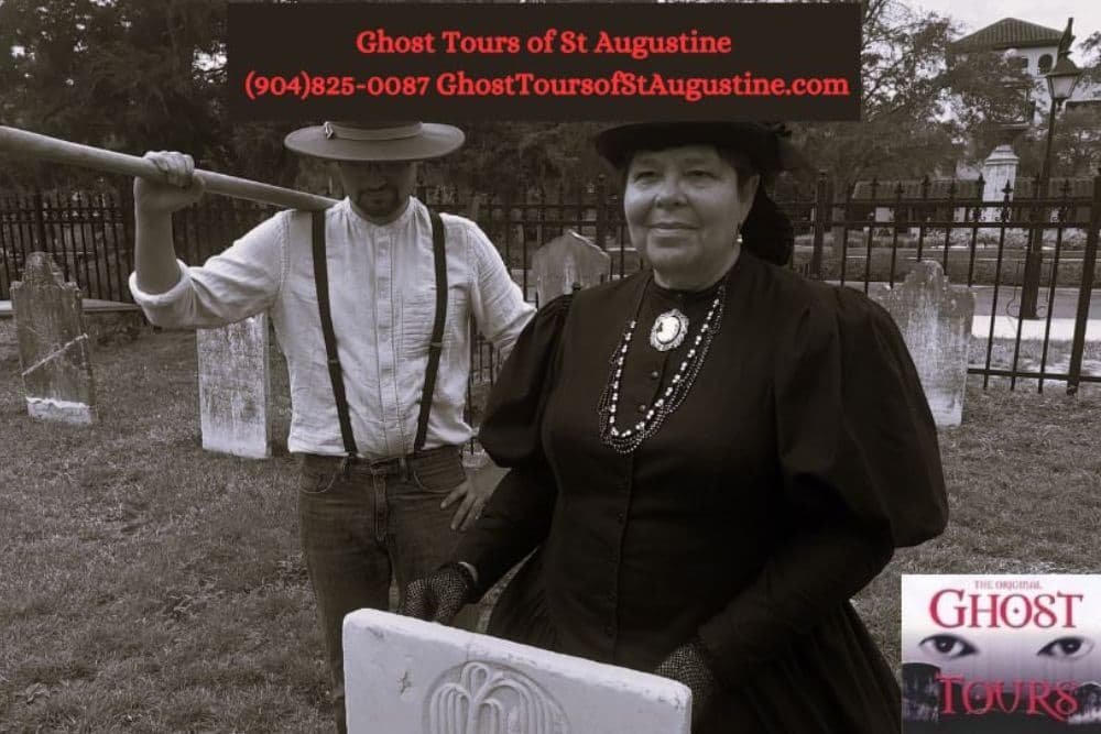 A woman dressed in historical attire stands in a graveyard while a man holds a shovel nearby.