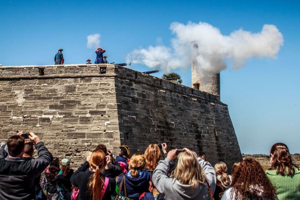 Tourists watch a cannon firing demonstration at a historic fort under a clear blue sky.