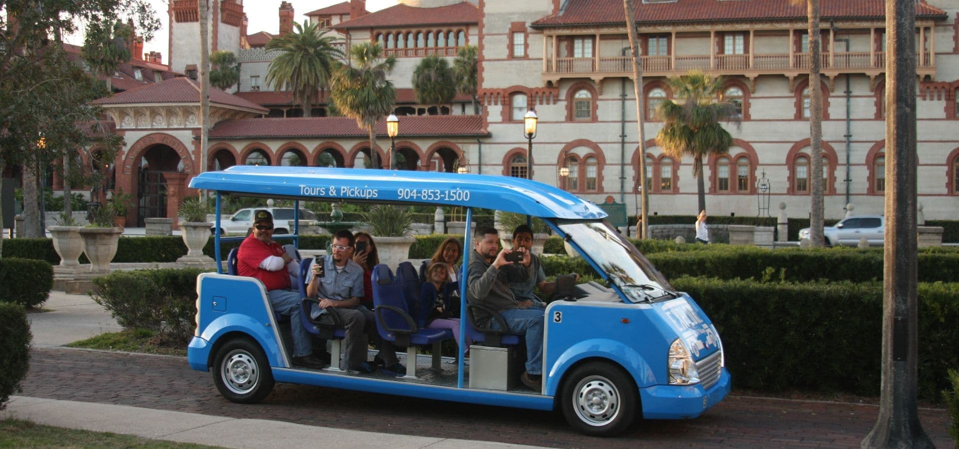 A blue tour vehicle with several passengers is parked in front of a historic building, surrounded by palm trees and greenery.