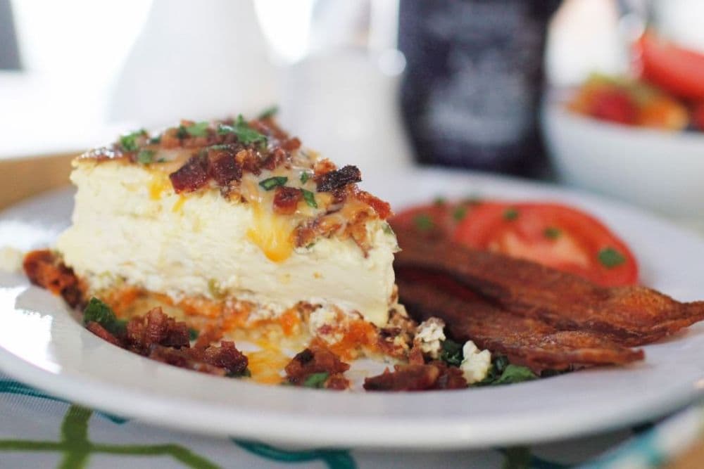 A slice of cheesy bacon egg casserole sits on a plate with crispy bacon and sliced tomatoes.