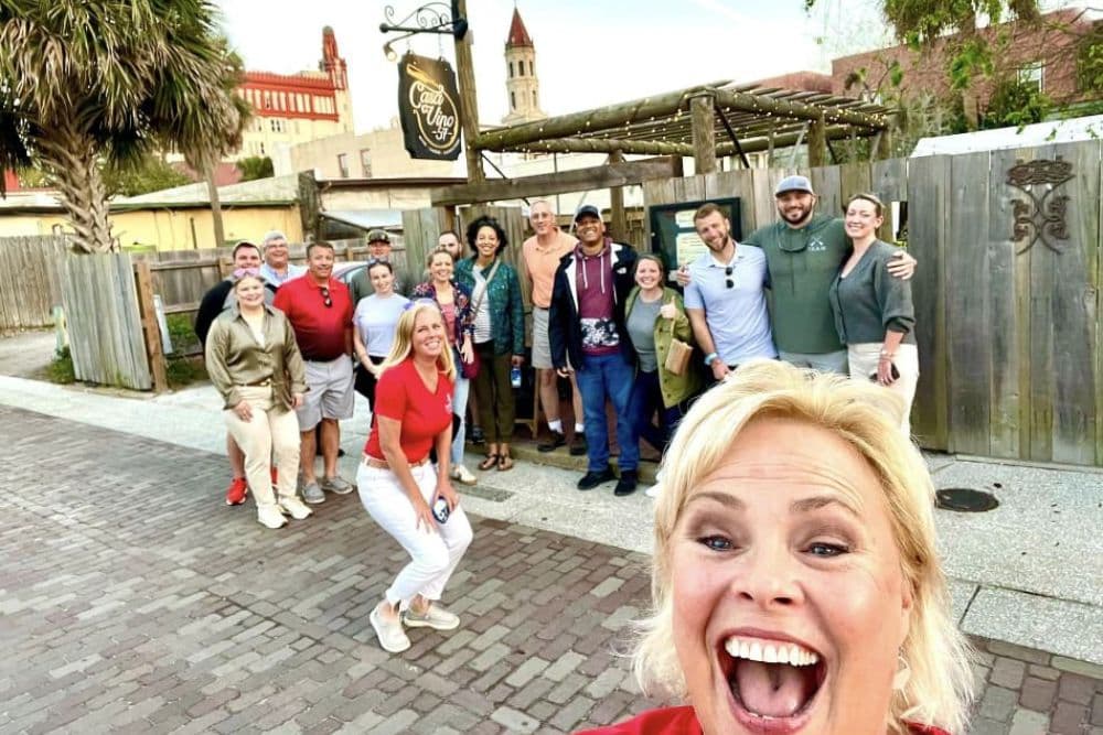 A group of people smiles together in front of a restaurant, with one person taking a selfie in the foreground.