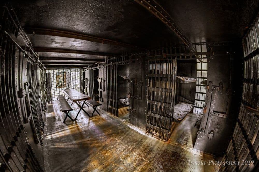 A dimly lit prison cell interior featuring barred doors, a long table, and simple bedding.