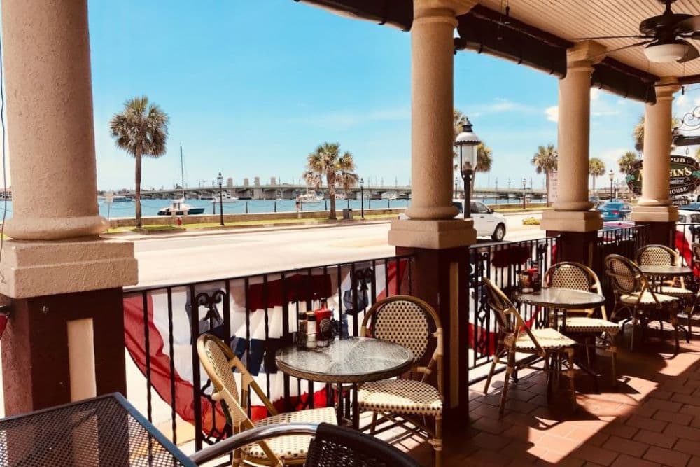 Outdoor seating area at a waterfront restaurant with a view of the bridge and palm trees.