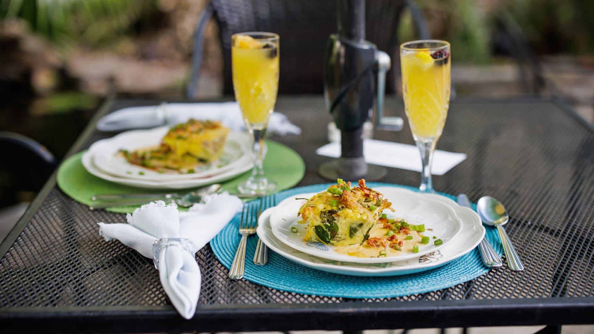 A beautifully arranged outdoor table with two plates of frittata and two glasses of mimosa.